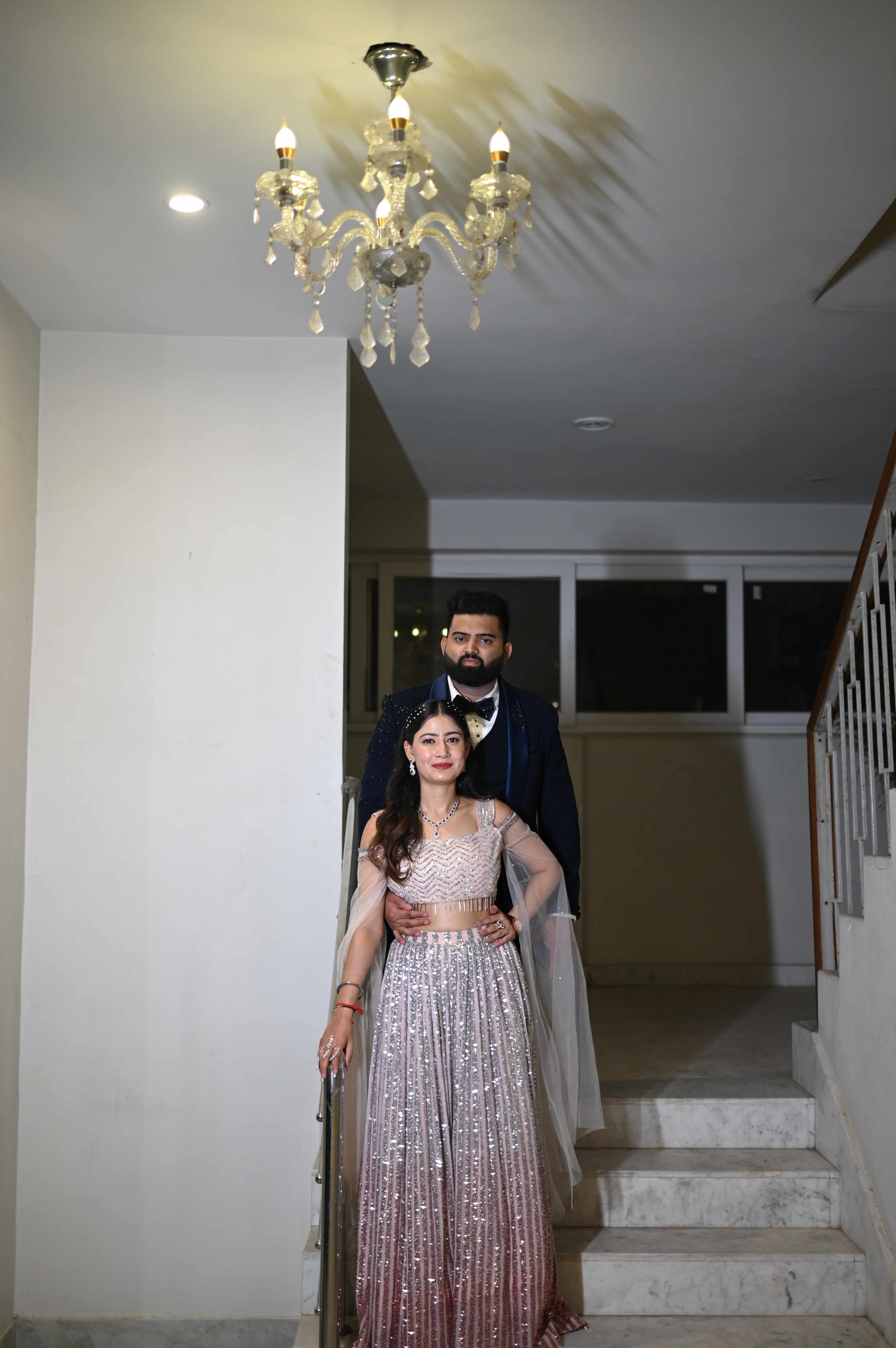 An elegantly dressed couple standing together on a staircase beneath a chandelier